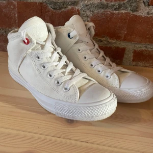 Vita Converse All Star sneakers - Säljer ett par klassiska vita Converse All Star sneakers i canvas med högt skaft. Skorna har snörning med en röd och en blå detalj i översta öljetterna, rund tå och platt sula i gummi. Perfekta för dig som gillar en clean och tidlös stil.