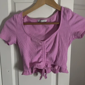 Ljuslila croppad topp från Zara - Supersöt ljuslila croppad topp från Zara med ribbad struktur, korta ärmar och snygg snörning framtill. Volangkant nedtill och knytdetalj ger en extra gullig touch. Perfekt för dig som gillar trendiga och färgglada plagg! Jättefin topp som jag köpte för 3 år sen inte kommit till användning efter det ☺️ Användt 4 gånger ‼️