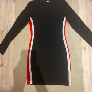 Svart bodycon klänning Divided H&M - Svart bodycon klänning från Divided H&M med långa ärmar och coola röda och vita ränder längs sidorna. Tight passform som framhäver figuren och sportig vibe. Perfekt för dig som gillar en enkel men ändå statement look.