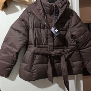 Brun pufferjacka i dun - Säljer en brun pufferjacka med huva och bälte i midjan. Jackan har quiltad design, dragkedja och långa ärmar. Perfekt för kalla dagar och har en snygg, oversized look. Säljes pga för stor