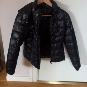 Svart pufferjacka från 157 XXS - Svart pufferjacka från 157 i storlek XXS. Jackan har glansig yta, horisontella quiltade sektioner och dragkedja framtill. Perfekt för kyliga dagar och har en klassisk look med hög krage och långa ärmar. Och luva 