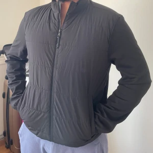 Svart pufferjacka med dragkedja - Svart pufferjacka med stilren design och hög krage. Jackan har quiltade paneler på framsidan och hel dragkedja. Materialet är syntet och polyester, vilket gör den lätt och bekväm. Perfekt för dig som gillar en clean look och vill ha något som funkar till många outfits.