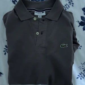 Brun pikétröja från Lacoste - Säljer en klassisk brun pikétröja från Lacoste med den ikoniska gröna krokodilloggan på bröstet. Tröjan har korta ärmar, krage och knäppning med två knappar. Tillverkad i bomull för en skön och luftig känsla. Perfekt för en clean och stilren look.