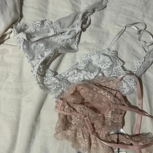 Spetsiga braletter från NA-KD - Två snygga braletter från NA-KD i spets, en i dusty pink och en i vitt. Båda har tunna axelband, justerbar knäppning och vacker blommig spets med volangkant. Perfekta att styla under en öppen skjorta eller kavaj för en trendig look öven en basic Bh! Säljer då dessa inte kommit till användning 