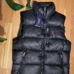 Svart dunväst  - Svart dunväst med hög krage och tre dragkedjeförsedda fickor. Klassisk röd logga broderad på bröstet. Västen har quiltad design och är perfekt för lager-på-lager. Snygg och stilren look med diskreta detaljer.