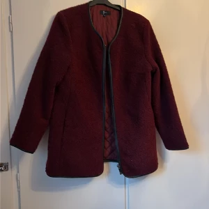 Vinröd teddyjacka från Geena - Säljer en vinröd teddyjacka från Geena i storlek 40. Jackan har långärmade ärmar, quiltat foder och dragkedja framtill. Materialet är mjukt och fluffigt, perfekt för höst och vinter. Enkel design med svarta kantdetaljer och rundad hals.