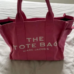 Rosa tote bag från Marc Jacobs - Säljer en rosa tote bag från Marc Jacobs med tydlig vit logga framtill. Väskan är i canvasmaterial, har två handtag och dragkedja upptill. Rymlig insida med innerficka, perfekt för att bära med sig allt man behöver. Snygg och trendig accessoar för alla tillfällen.