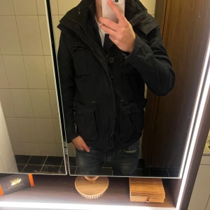 Svart parkajacka Abercrombie & Fitch - Svart parkajacka från Abercrombie & Fitch med huva och flera stora fickor framtill. Jackan har snörning i midjan och knappar samt dragkedja. Snygg och funktionell design, perfekt för dig som gillar streetstyle och vill ha en jacka med coola detaljer.