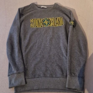 Grå Stone Island sweatshirt M - Säljer en grå sweatshirt från Stone Island i storlek M. Tröjan har broderad logga och text i gult och svart framtill samt klassisk patch på ärmen. Tillverkad i mjuk bomull med ribbade muddar vid ärmslut och nederkant. Perfekt för en avslappnad streetwear-look.