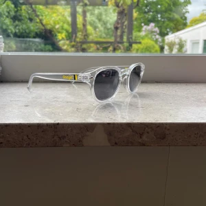 Gnaget solglasögon med transparent båge ( Limited edition) - Snygga solglasögon med transparent rundad båge och mörkgrå linser. På skalmen finns en gul och svart detalj med texten 'Gnaget'. Perfekt för dig som vill sticka ut med en cool och modern stil.