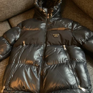 Svart pufferjacka Alessandro Milano - Snygg svart pufferjacka från Alessandro Milano med glansig finish och huva. Jackan har flera dragkedjor framtill, en badge på bröstet och paneler i matt tyg. Perfekt för kalla dagar och har en modern, urban vibe.