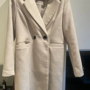 Beige kappa med knappar - Snygg beige kappa med klassisk krage och dubbelknäppning framtill. Jackan har två stora fickor och är helfodrad för extra komfort. Långärmad och med stilren, rak passform som ger en clean look. Perfekt för dig som gillar minimalistisk stil.