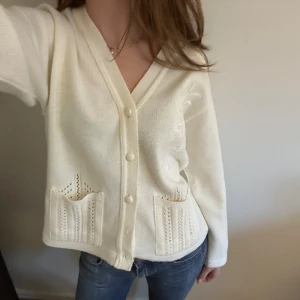 Vit kofta med broderi och fickor - En vit kofta med v-ringning, dekorativa broderier på bröstet och två stickade fickor framtill. Koftan har knappar hela vägen och är långärmad. 