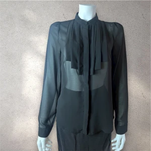 Svart transparent blus med knyt - Svart transparent blusf rån Vila  med lång ärm och snygga knytband framtill. Blusen har en liten ståkrage med knapp och är tillverkad i ett lätt, flödande material som ger en cool och elegant vibe. Perfekt för dig som gillar stilrena detaljer och vill sticka ut. #vila #blus