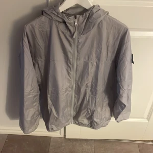 Ljusgrå vindjacka från Stone Island - Snygg ljusgrå vindjacka från Stone Island med huva och dragkedja framtill. Sitter lite för litet men den är helt ny och oanvänd.