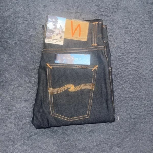 Nudie Jeans Grim Tim  - Helt nya jeans från Nudie Jeans, modellen Grim Tim med slim straight leg. Klassisk femficksdesign, orange kontrastsömmar och läderpatch med logga bak. Jeansen är tillverkade i slitstarkt denim och har en stilren look som passar perfekt till sneakers. Midja 34cm, längd 110cm.  Ifall det inte finns någon bild i annonsen där jag bär plaggen jag säljer så är det för att jag ej kan ha storleken heller passformen, så kolla måtten för storlek och googla på modellnamnet för passform 😉44