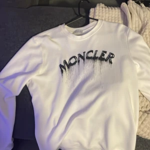 Vit sweatshirt från Moncler.  S/M - Säljer en clean vit sweatshirt från Moncler med svart drip-printad logga framtill och klassisk broderad Moncler-patch på ärmen. Tröjan har rund hals och är långärmad, perfekt för dig som gillar streetwear och exklusiva märken.