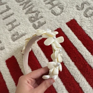 Diadem med blommor och pärlor - Ett superfint diadem i ljus cremevitt med stora blommor i satinliknande material och pärldetaljer i mitten av varje blomma. Perfekt accessoar för att lyfta vilken outfit som helst med en söt och elegant touch.