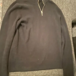 Svart stickad tröja med half zip - En svart stickad tröja med lång ärm och half zip-dragkedja vid halsen. Enkel och stilren design som passar perfekt till lager-på-lager under höst och vinter. Tröjan har en rak passform och är mjuk och skön att bära.