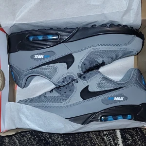 Nike Air Max 90 grå sneakers - Nike Air Max 90 sneakers i grått med svarta och blå detaljer. Skorna har en klassisk siluett med synlig Air-enhet i sulan, mesh och syntetmaterial på ovandelen, samt snörning och rund tå. Snyggt Nike Air-tryck i blått på hälen och Air Max-logga på sidan. Skickar med skolåda.