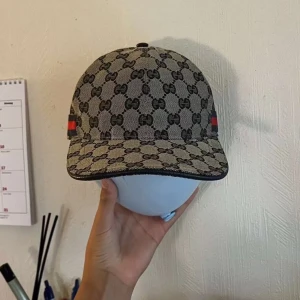 Gucci keps med GG-mönster och rödblå rand  - Snygg keps från Gucci i beige och marinblått med klassiskt GG-monogram över hela kepsen. På sidan finns en ikonisk röd och marinblå rand. Kepsen är tillverkad i bomullscanvas och har detaljer i mörkblått tyg på insidan. Perfekt för dig som vill sticka ut med lyxig streetstyle. Helt ny och oanvänd pris går att sänkas och diskutera