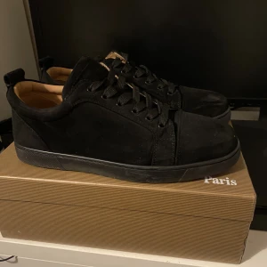Svarta sneakers från Christian Louboutin - Säljer ett par svarta sneakers från Christian Louboutin med klassisk röd sula och snygg signatur. Skorna har rund tå, snörning och är tillverkade i mocka med detaljer i skinn. Perfekt för dig som vill ha en lyxig och stilren look.