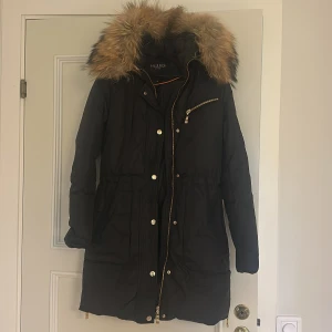 Svart parkas med päls från Hollies - Svart lång jacka från Hollies. Kemtvättad. Snygg päls på huvan och guldfärgade detaljer. Jackan har dragkedja och knappar framtill samt en praktisk bröstficka med dragkedja. Superfin och varm – perfekt för kalla dagar med sin stilrena look.  Obs: Jackan sprack lite på höger axel när en tjej provade den, men det går att sy utan problem, därför är priset lägre. Annars är jackan i jättebra skick.