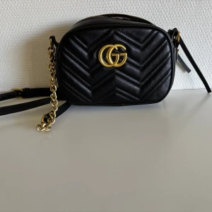 Svart axelväska  - Snygg svart axelväska med guldiga detaljer och logga framtill. Väskan är tillverkad i skinn och har ett quiltat mönster med dragkedja upptill. Kedja och läderrem ger en cool vibe och den kompakta formen gör den perfekt för det viktigaste. Tyvärr har väskan slitage.