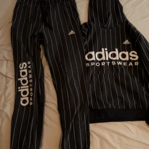 Svartrandig tracksuit från Adidas - Svarta mjukisbyxor från Adidas med vita vertikala ränder och stor Adidas Sportswear-logga längs benet. Byxorna har elastisk midja, bakficka och är gjorda i mjukt bomullsmaterial. Perfekt för chill eller sportiga dagar. Hoodien har lös passform, stor ficka på magen och stor logga. Katt finns i hemmet. 