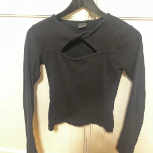 Svart ribbad topp med cut out Gina Tricot - Svart långärmad topp från Gina Tricot med ribbad struktur och snygg cut out-detalj vid halsen. Toppen har en tight passform och cool twistad design upptill som ger en unik look. Perfekt för dig som vill sticka ut lite extra.