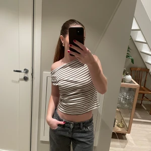 Randig offshoulder topp  - Säljer en vit och brunrandig offshoulder topp i croppad modell. Toppen är ärmlös och har en asymmetrisk, oneshoulder-skärning som ger en snygg och trendig look. Materialet är mjukt och stretchigt. Perfekt till jeans eller kjol för en chill stil.