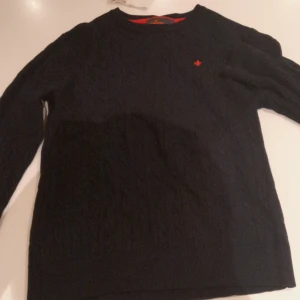 Svart kabelstickad tröja Morris L - Svart kabelstickad tröja från Morris i 100% merinoull. Klassisk modell med rund halsringning och diskret röd logga broderad på bröstet. Perfekt för dig som gillar stilrena och tidlösa plagg med premiumkänsla.