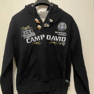 Svart hoodie Camp David - Svart hoodie från Camp David med broderade detaljer och tryck både fram och bak. Tröjan har huva med snörning. Snygga marinblå och vita brodyrer.