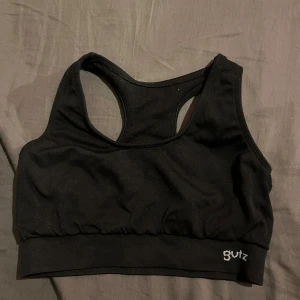 Svart sportig linnetopp från Gutz - Svart sportig linnetopp från Gutz med brottarrygg och bred resår nedtill. Perfekt för träning och har en stilren design utan mönster. Materialet känns mjukt och stretchigt, vilket ger bra komfort och stöd under aktivitet.