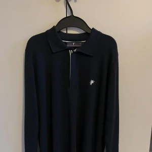 Marinblå pikétröja från Denim Culture - Snygg marinblå pikétröja från Denim Culture med lång ärm och half zip-dragkedja framtill. Tröjan har ett diskret mönster i tyget och vit broderad logga på bröstet. Kragen är klassisk och dragkedjan har vita detaljer som matchar loggan. Hör av er vid funderingar💫