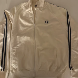 Vit Fred Perry tröja  - Snygg vit track jacket från Fred Perry med marinblå ränder längs ärmarna och broderad logga på bröstet. Jackan har ribbad krage, muddar och dragkedja framtill. Perfekt för en sportig och clean stil. Liten i storleken så passar L men även som större M. Pris kan diskuteras vid snabb affär 