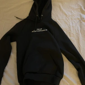 Svart hoodie Peak Performance S - Svart hoodie från Peak Performance i storlek S. Klassisk design med vit logga på bröstet, huva med snörning och stor magficka. Mjuk och skön bomullsmix, perfekt för chill eller träning. Långärmad och med ribbade muddar för extra komfort.