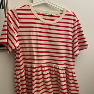 Randig röd och vit tröja från Zara - Superfin tröja från Zara i rött och vitt med horisontella ränder. Modellen har rund halsringning, korta ärmar och en lätt rynkad midja som ger en avslappnad och söt siluett. Tillverkad i mjuk bomull, perfekt för varmare dagar.