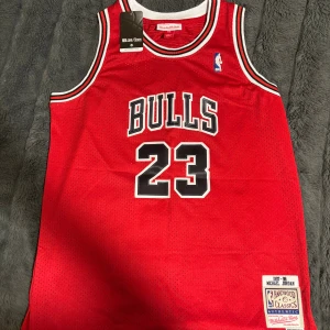 Chicago Bulls Jordan 23 baskettröja - Klassisk Chicago Bulls baskettröja från Mitchell & Ness, Michael Jordan 23, röd med svarta och vita detaljer. Broderade loggor och namn, meshmaterial för extra ventilation. Snygga kontrastkanter vid hals och ärmhål. 