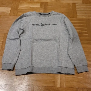 Grå Sail Racing sweatshirt i bomull - Snygg grå sweatshirt från Sail Racing med svart logotyp tryckt på bröstet. Tröjan har rund hals, ribbade muddar och lång ärm. Tillverkad i mjuk bomullsmix som är skön att ha på sig. Perfekt för dig som gillar stilren och sportig look.