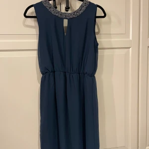 Mörkblå klänning med glittrig krage - Supersnygg mörkblå klänning med glittrig, dekorerad krage och ärmlös design. Klänningen har en markerad midja med resår och ett litet nyckelhålsutskärning fram. Perfekt för dig som vill sticka ut med lite extra bling.