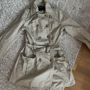 Beige trenchcoat från Reporter S - Snygg beige trenchcoat från Reporter med klassisk dubbelknäppning, stora fickor och bälte i midjan. Jackan har långa ärmar, bred krage och coola detaljer som axelklaffar och stora knappar. Perfekt för dig som gillar stilren och tidlös look.