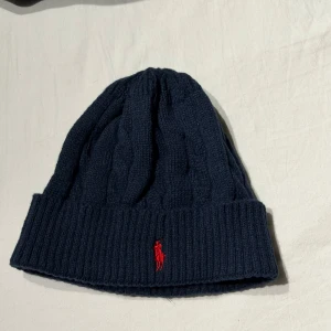 Mörkblå kabelstickad mössa Polo Ralph Lauren - Snygg mörkblå kabelstickad mössa från Polo Ralph Lauren med röd broderad logga framtill. Mössan har bred uppvikt kant och är tillverkad i mjuk ull som håller dig varm. Perfekt accessoar för kyliga dagar med klassisk stil.