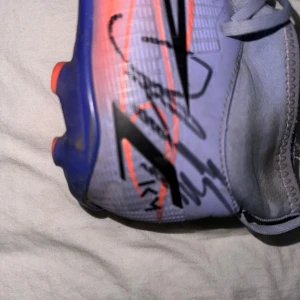 Lila och orange fotbollssko med autograf - Säljer en Nike fotbollssko i lila och orange med svarta detaljer och snörning. Skon har en platt sula med dobbar för gräsplan och är tillverkad i syntetmaterial. Sidan pryds av en autograf, vilket gör skon unik och extra cool för samlare eller fotbollsfans. Autografen är skriven av Tottenham och Sverige national lag i fotboll, dejan kulusevski 