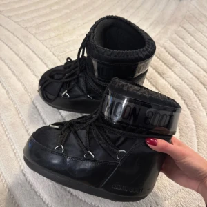 Svarta Moon Boots - Moon Boots i helsvart med glansig yta och tydlig logga runt skaftet. Skorna har snörning framtill och en rund tå, samt tjock platt sula som ger extra komfort. Perfekta för kalla vinterdagar och har en unik, futuristisk look som sticker ut.