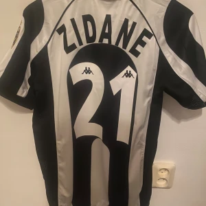 Juventus Zidane #21 fotbollströja  - Juventus fotbollströja med ZIDANE och nummer 21 på ryggen. Svartvit randig design, klassisk krage och korta ärmar. Kappa-logga på bröstet och axlarna, Champions League-märke på ena ärmen och Juventus klubbmärke på andra. Materialet är polyester. Tagg finns kvar!