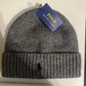 Grå ribbstickad mössa Polo Ralph Lauren - Snygg grå ribbstickad mössa från Polo Ralph Lauren med klassisk uppvikt kant och broderad svart logga framtill. Tillverkad i mjukt ullmaterial som håller dig varm under kyliga dagar. Perfekt accessoar för en stilren look.