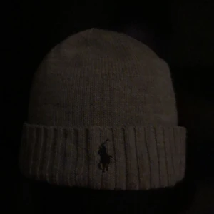Grå stickad mössa Polo Ralph Lauren - Snygg grå stickad mössa från Polo Ralph Lauren med klassisk ribbad kant och broderad logga framtill. Perfekt accessoar för kyliga dagar och enkel att matcha med olika outfits.