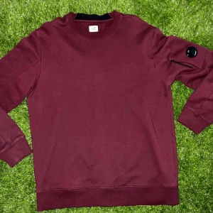 Vinröd sweatshirt från C.P. Company XL - Säljer en vinröd sweatshirt från C.P. Company i storlek XL. Tröjan har lång ärm, rund hals och en ikonisk svart linsdetalj med dragkedja på vänster ärm. Materialet är mjuk bomull och passformen är normal. Perfekt för dig som gillar streetwear och snygga detaljer.