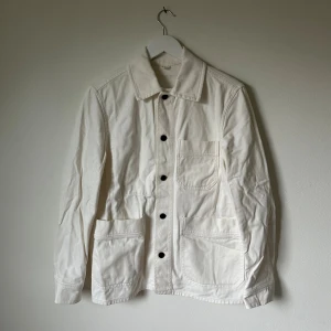 Vit overshirt från Arket, storlek 46 - Snygg vit overshirt från Arket i bomullstwill. Jackan har klassisk krage, svarta knappar framtill och tre stora fickor på framsidan. Perfekt för lager-på-lager och enkel att matcha med jeans eller chinos. Långärmad och stilren design. 4B5